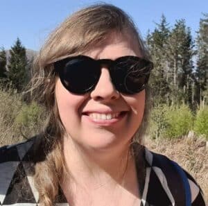 Photo of Jacq the admin wearing sunglasses, outside with trees in background and blue skies.