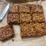 Vegan chocolate flapjacks with one cut off from main batch, knife to side, baking parchment underneath flapjack.