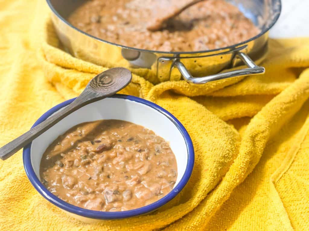 Plant-based vegan refried beans in small white enamel bowl with blue rim, wooden spoon, and cooking skillet to side, all sitting on yellow tea towel.