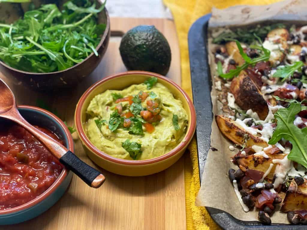 vegan loaded black bean potato wedges with guacamole dip, salsa and salad in bowl, avocado on wooden board.