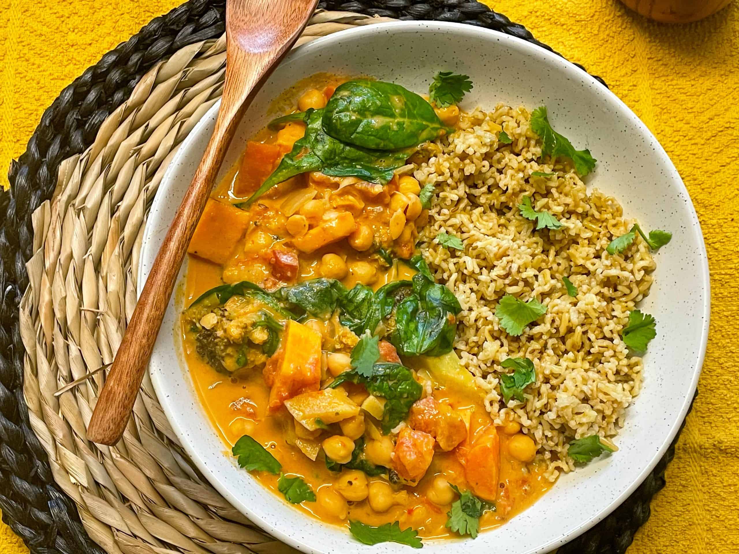 A cream bowl of Thai red curry full of veggies, wooden spoon to side, wicket mat underneath.