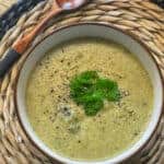 Cream of kale and celery soup in a brown rimmed bowl with a basket style placemat and dark wooden spoon to side, yellow tablecloth, featured image.