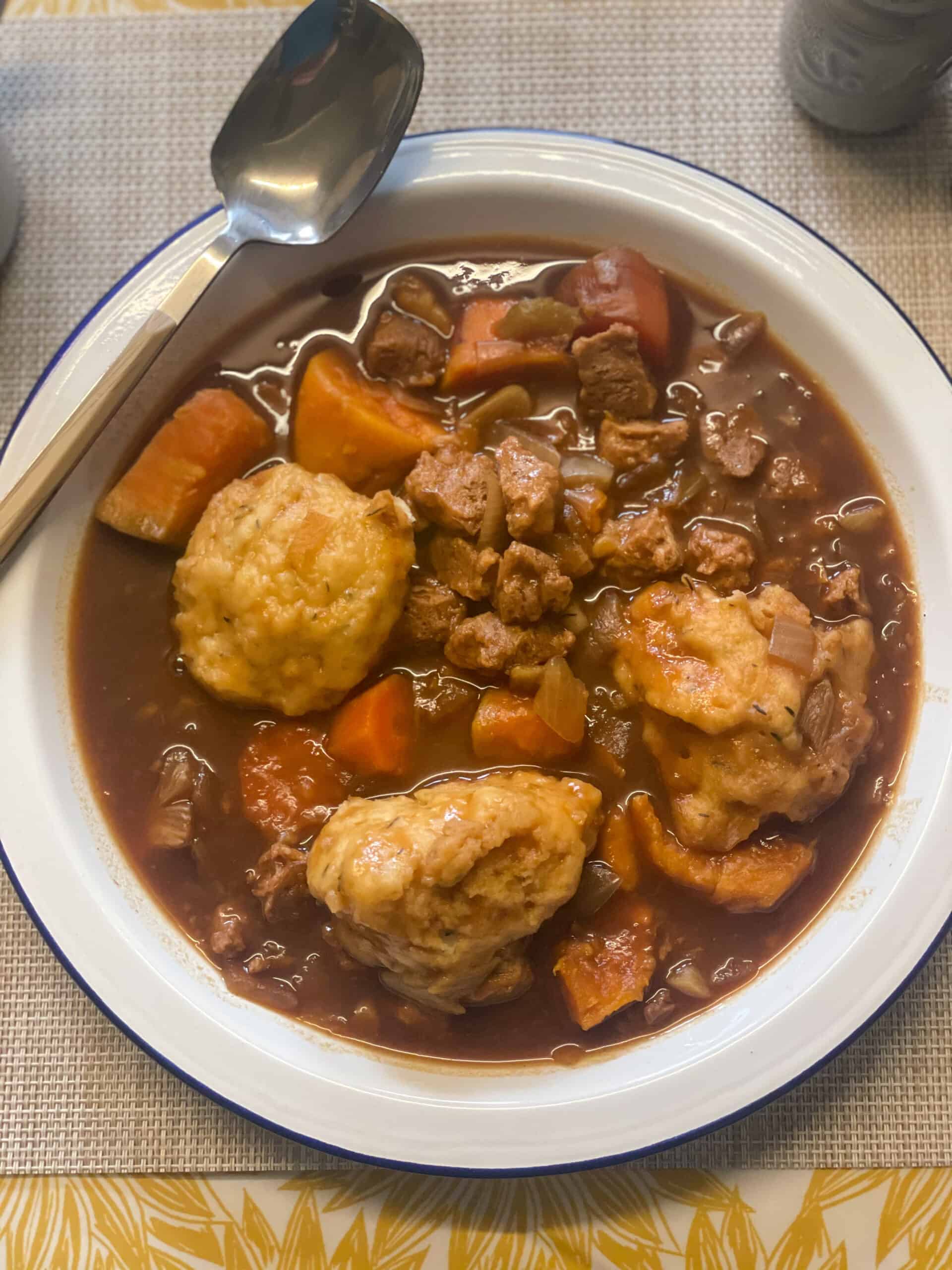 slow cooker vegan beef stew and dumplings ready to eat in white bowl with blue rim, spoon at side of plate