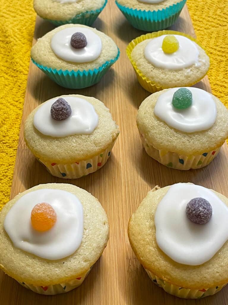  fairy cakes decorated with water icing and a jelly tot sweet, wooden long chopping board background.