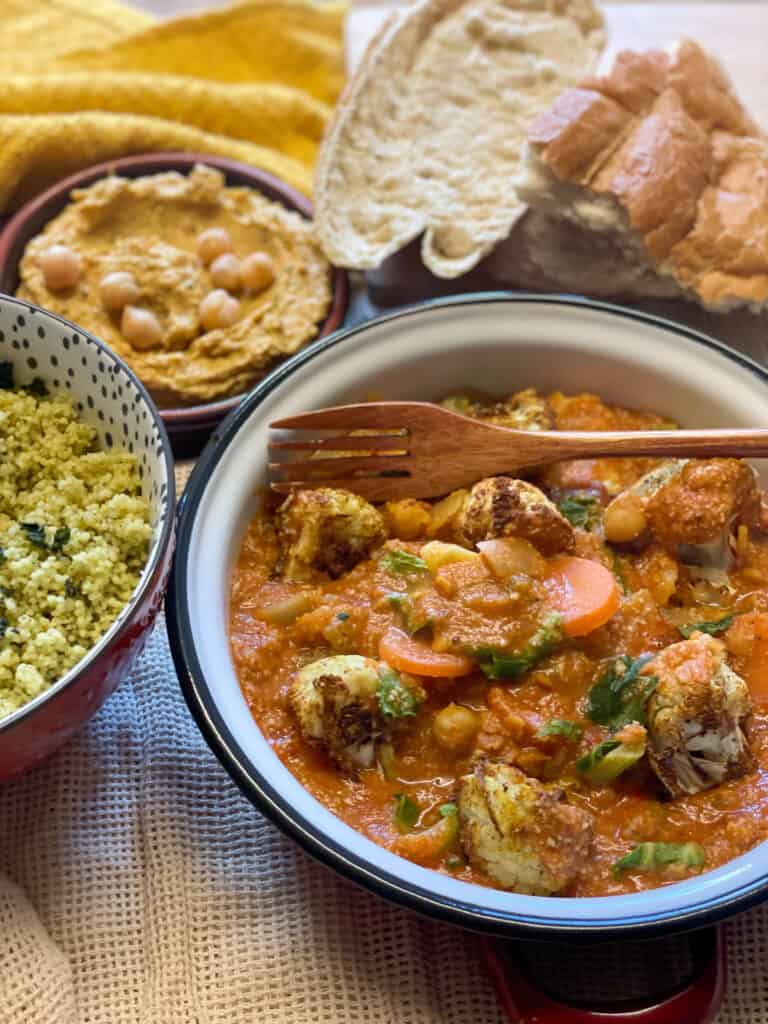 Moroccan stew served in a bowl with bowl of cous cous to side, a bowl of hummus to side and wedges of crusty bread.