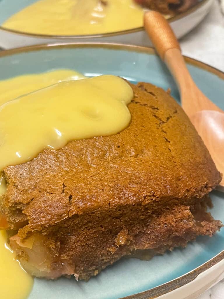 Pudding added to serving dish, with custard poured over, wooden eating spoon to side.