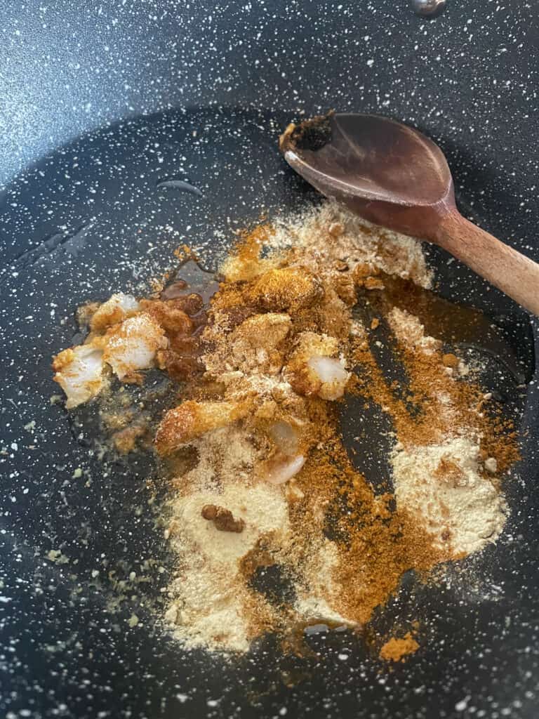 Curry powder and garlic cooking in the skillet with a wooden spoon at the side of pan.