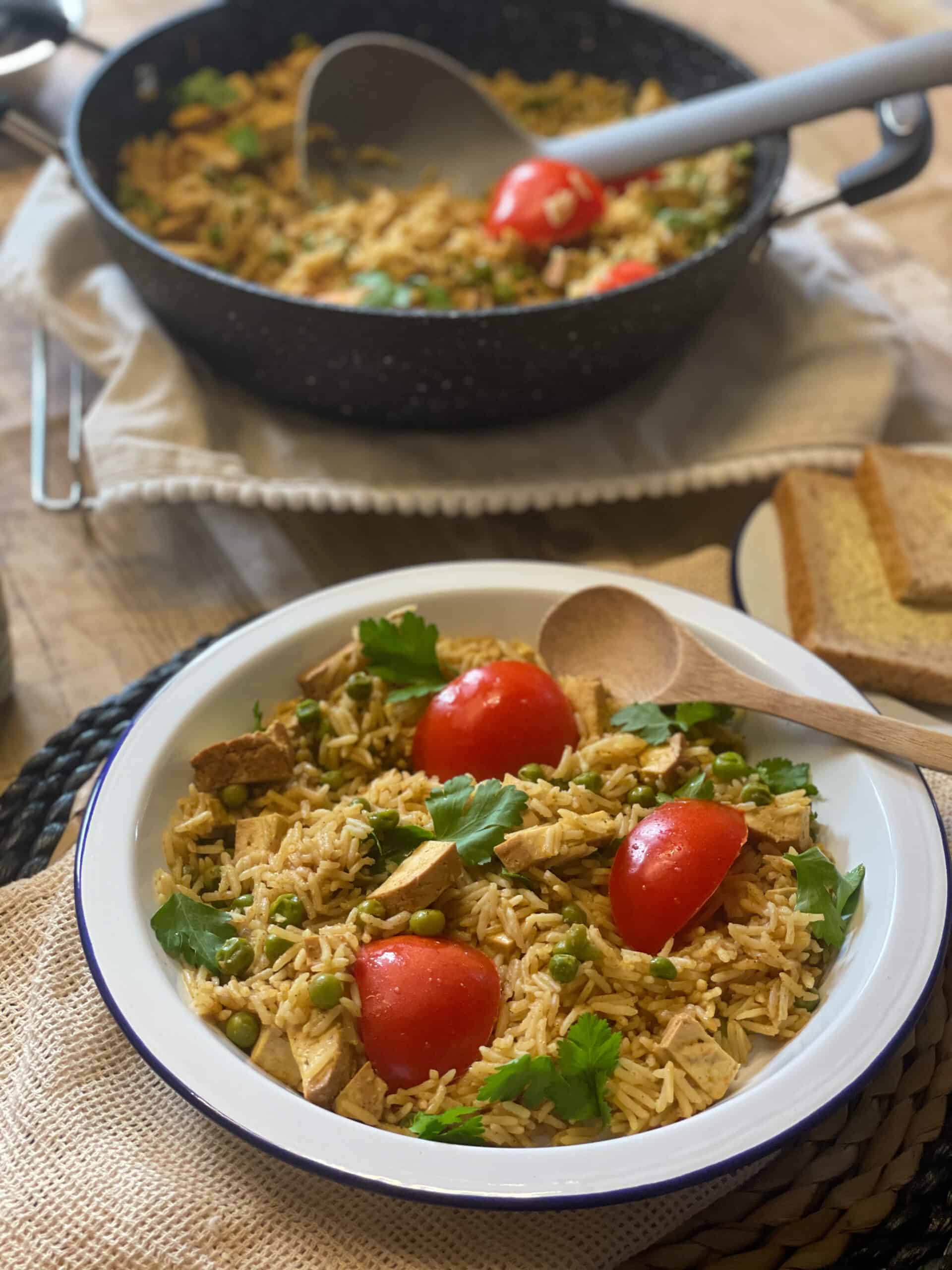 Vegan Kedgeree served in a white bowl with a dark coloured pan in background, cream coloured table cloth.