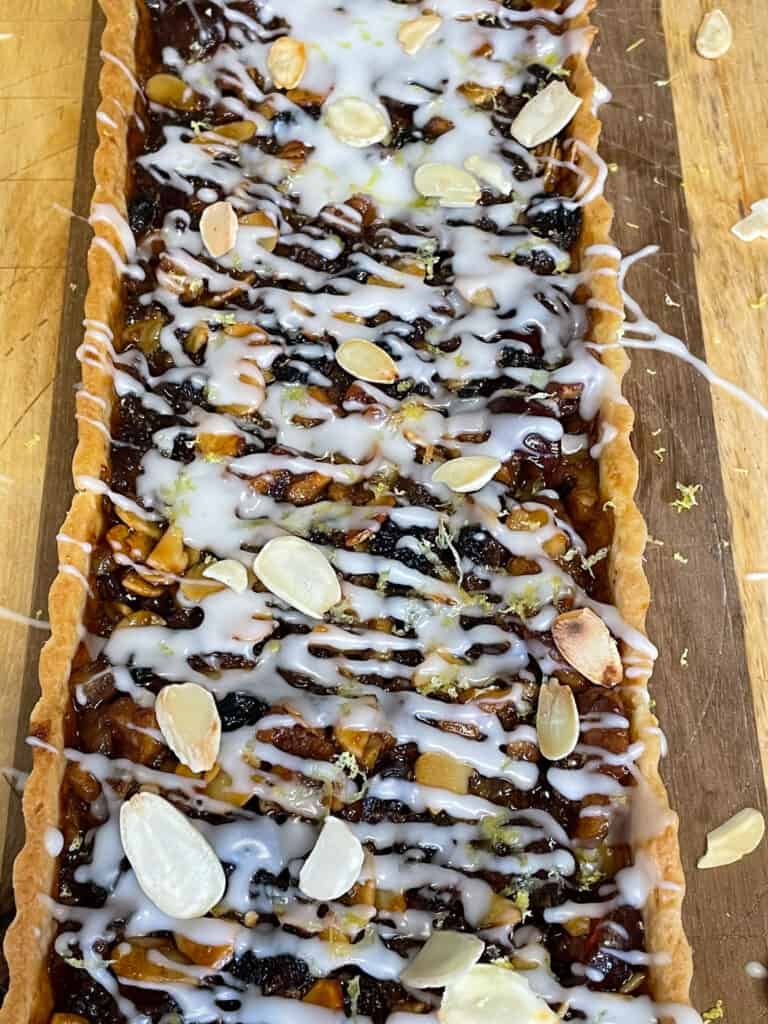 Close up of rectangular vegan Scottish Border tart unsliced on wooden chopping board with flaked almonds and lemon zest scattered around.
