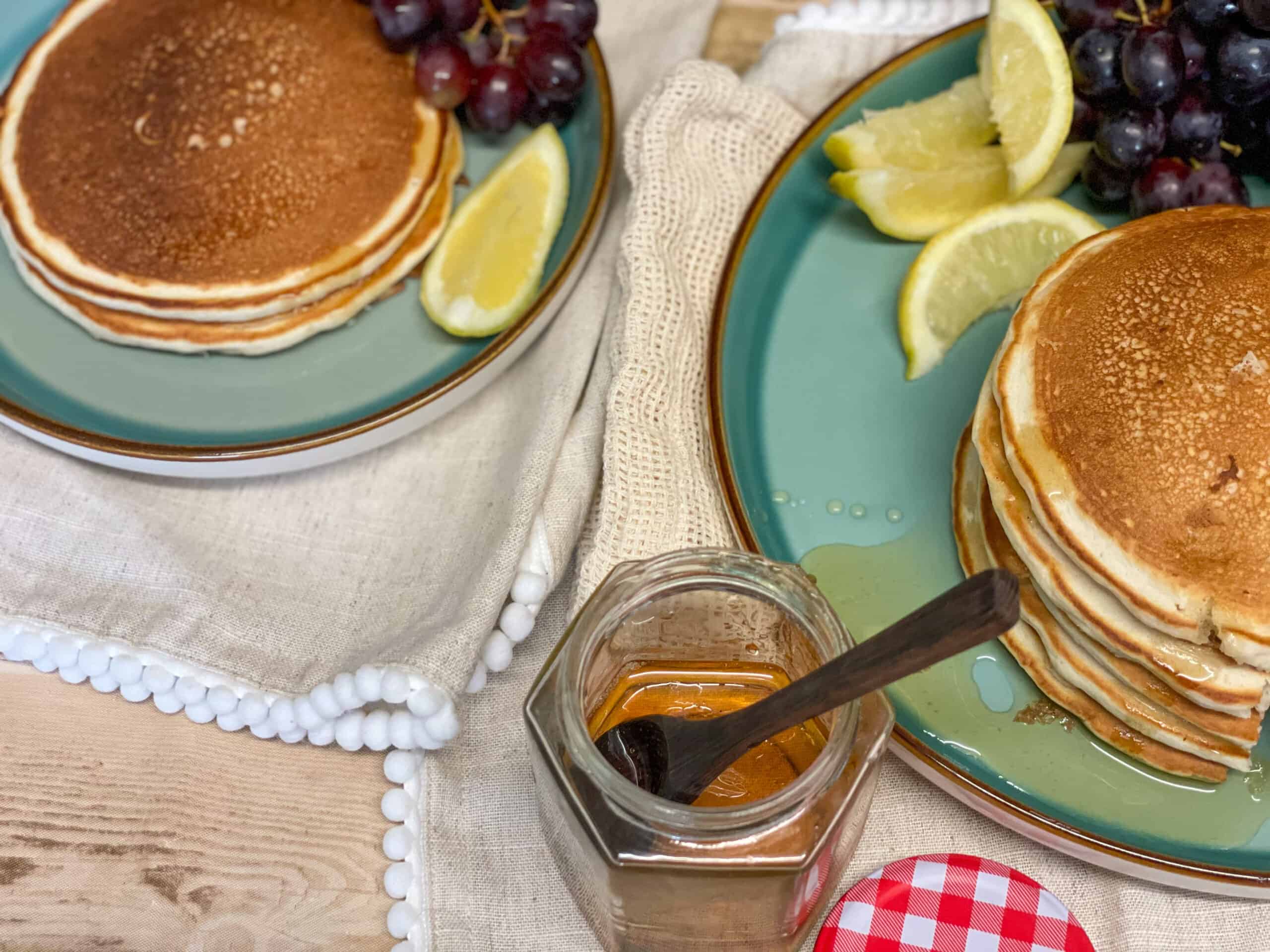 vegan traditional Scotch pancakes served with dandelion honey in a jar with small wooden spoon, lemon slices and grapes to side.