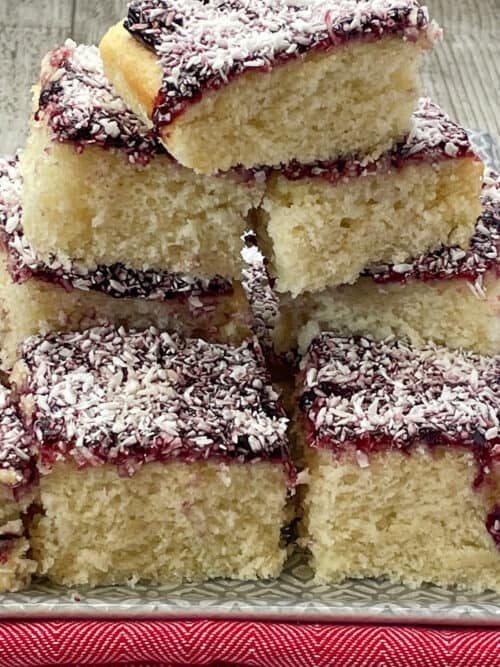 A pile up of vegan sponge cake with a jam and coconut topping, with a light grey background.