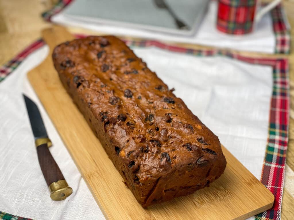 vegan fruit loaf cake sitting on a chopping board with tartan napkin and brown handled knife