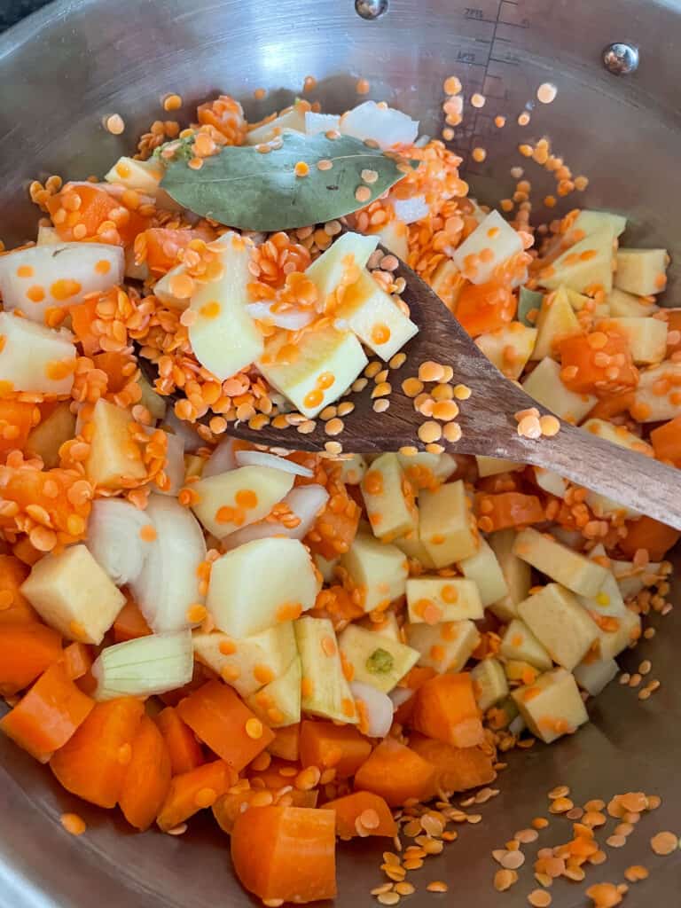 diced potato, carrot, swede, onion, and red lentils, bay leaf and stock cubes in a saucepan, with wooden mixing spoon