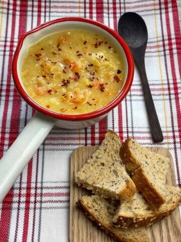 Vegan lentil soup in a small enamel cream pan with a red rim, wooden spoon, small board off to the side with pieces of bread, and a check red and white tea towel underneath.