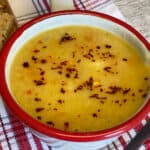 Lentil soup with red pepper flakes garnish, pieces of brown bread, wooden spoon to side and red check tea towel underneath.