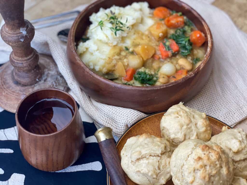 potage stew in a brown bowl with little bread rolls on a plate, with a brown handled knife at the side, small brown wooden cup with water and a wooden candle stick with wax drips. 