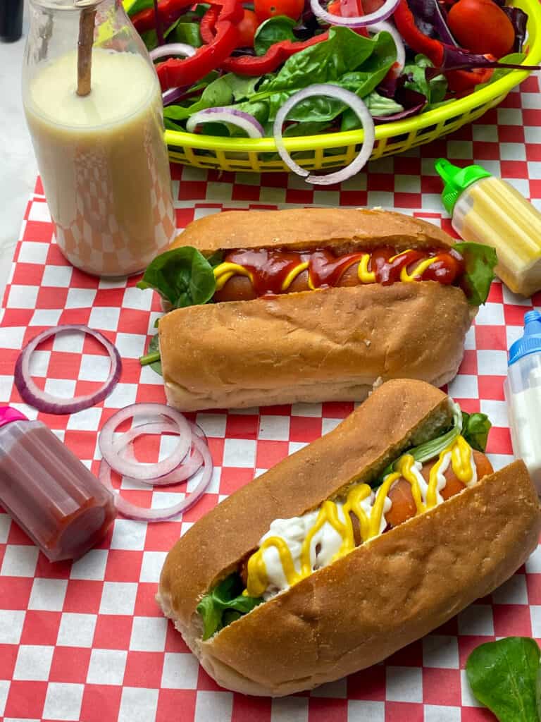 carrot hotdogs, with small glass bottle with milkshake and straw, small sauce bottles and a basket of mixed salad, on top of a red check mat