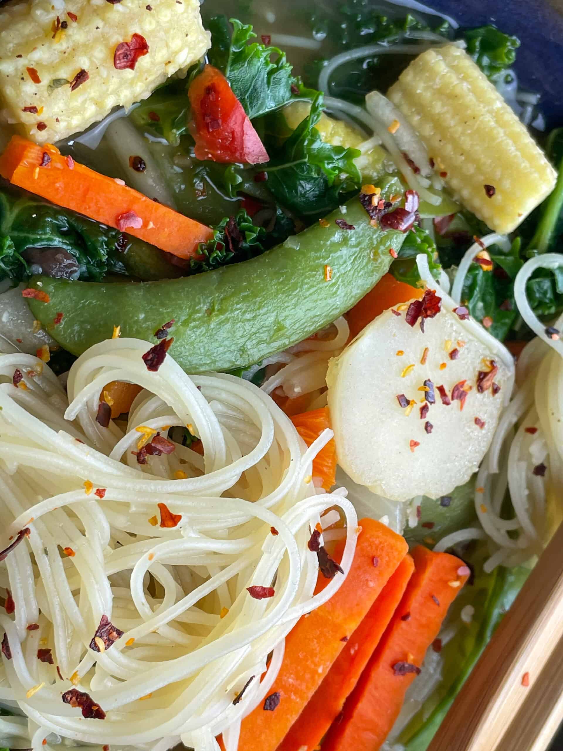 close up of vegan stir fry soup, noodles, peapod, mini corn cobs, carrot batons and chop stick to side.