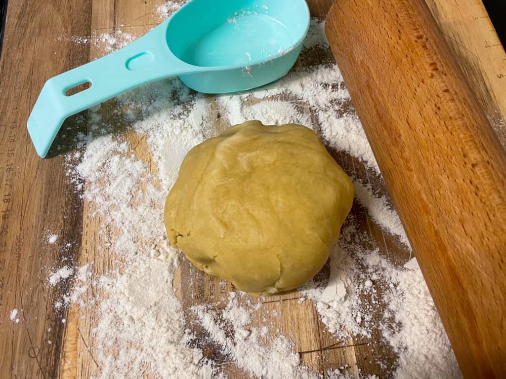 Pastry ball, with measuring cup and wooden spoon to side, flour sprinkled on chopping board.