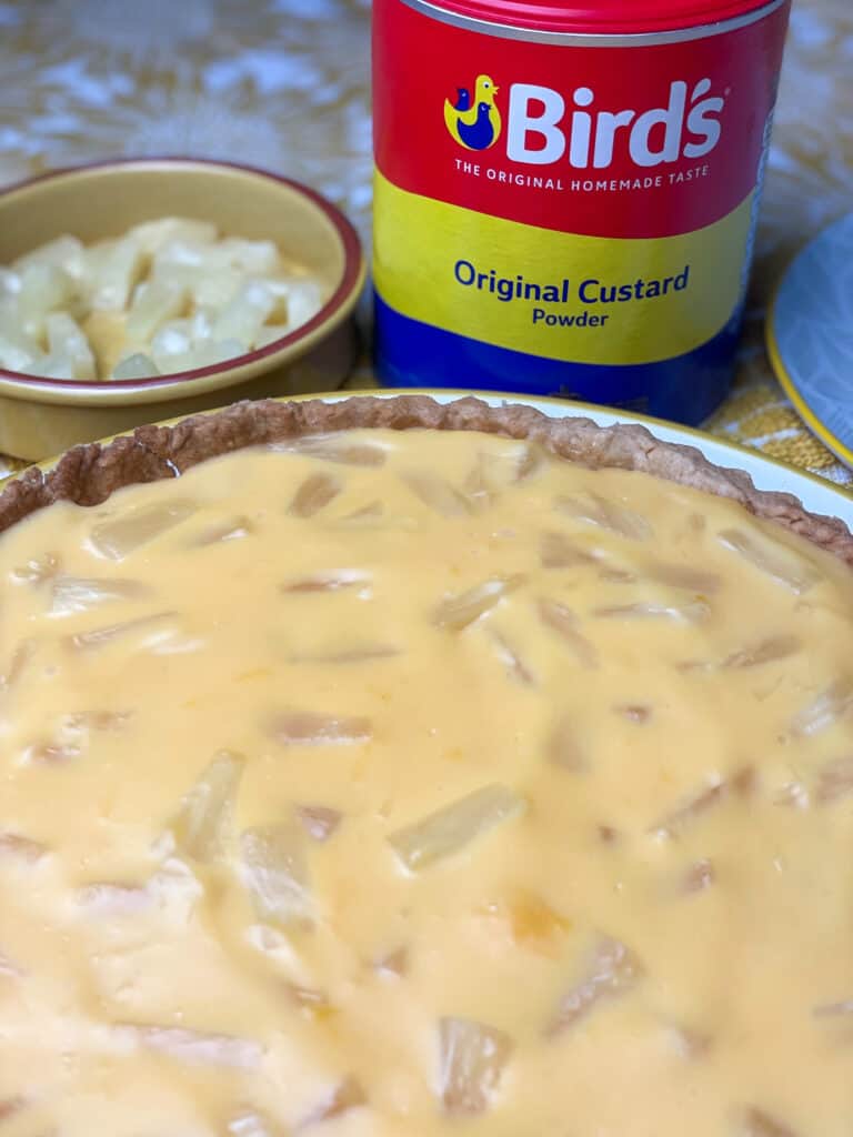 custard tart with Birds custard powder tin in background, and small dish with chopped pineapple pieces.