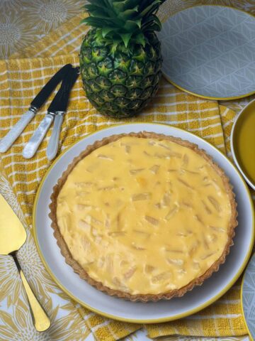 Vegan pineapple tart on serving plate, with dessert knifes off to side, serving plates at side, serving spatula to side and a green fresh pineapple at top, featured image.