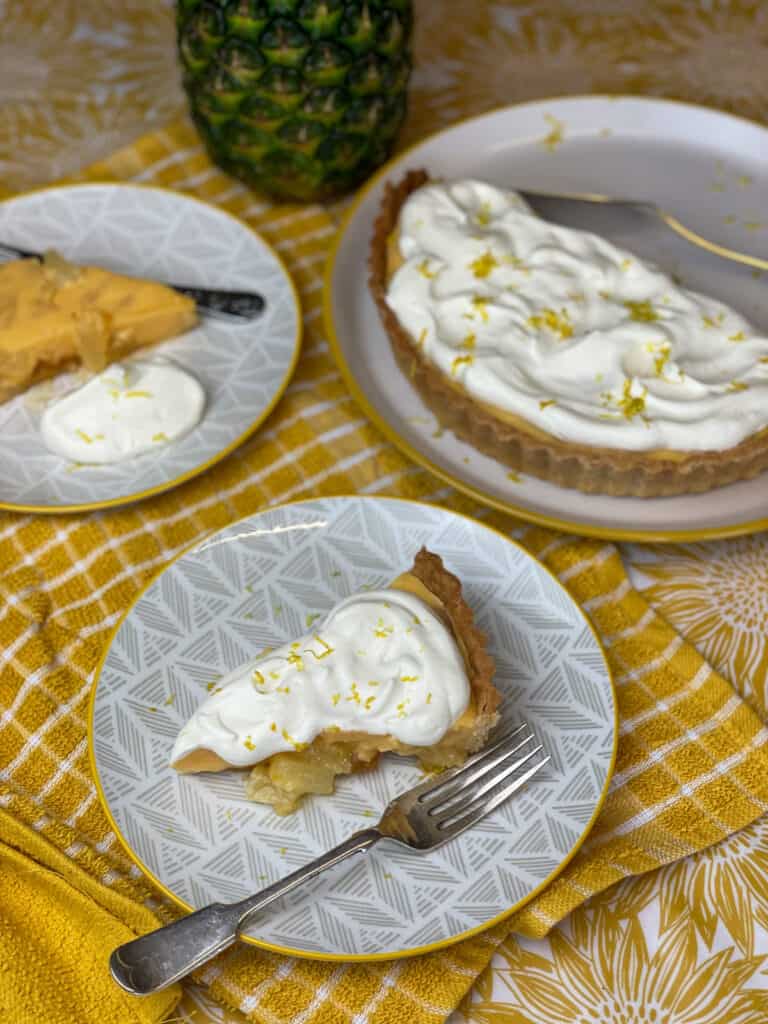 pineapple tart with whipped cream topping, 3 plates with large tart, and 2 smaller pieces. Yellow check tea towel background.