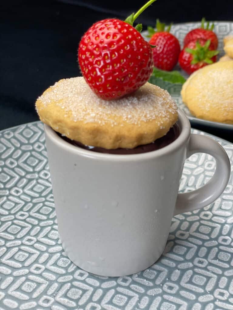 close up of chocolate pudding in grey expresso mug with shortbread biscuit on top and strawberry, black background and grey plate.