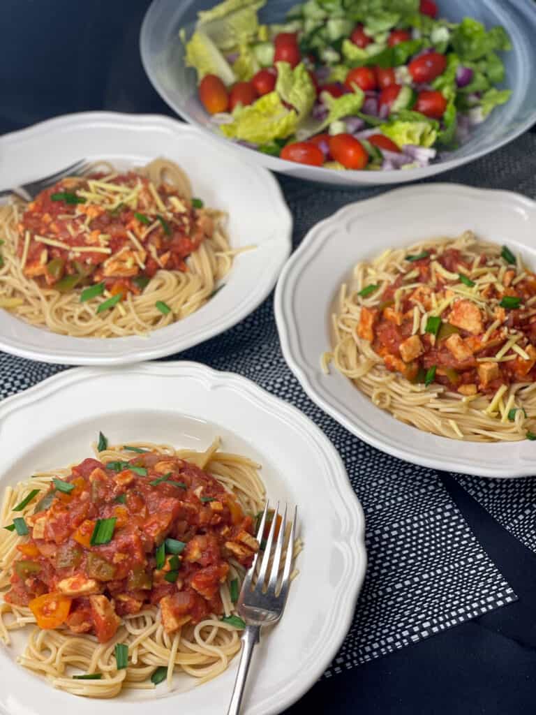 3 bowls of tempeh Bolognese with chopped chives and a salad bowl in background.