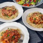 3 bowls of tempeh Bolognese with chopped chives and a salad bowl in background.