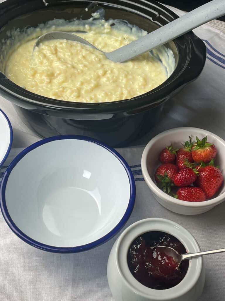 rice pudding in slow cooker with empty serving bowl, fresh strawberries and strawberry jam pot.