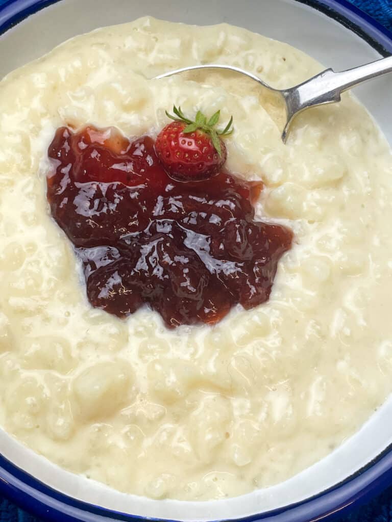 Close up of rice pudding with strawberry jam and wee fresh strawberry in white bowl with blue rim and silver spoon.