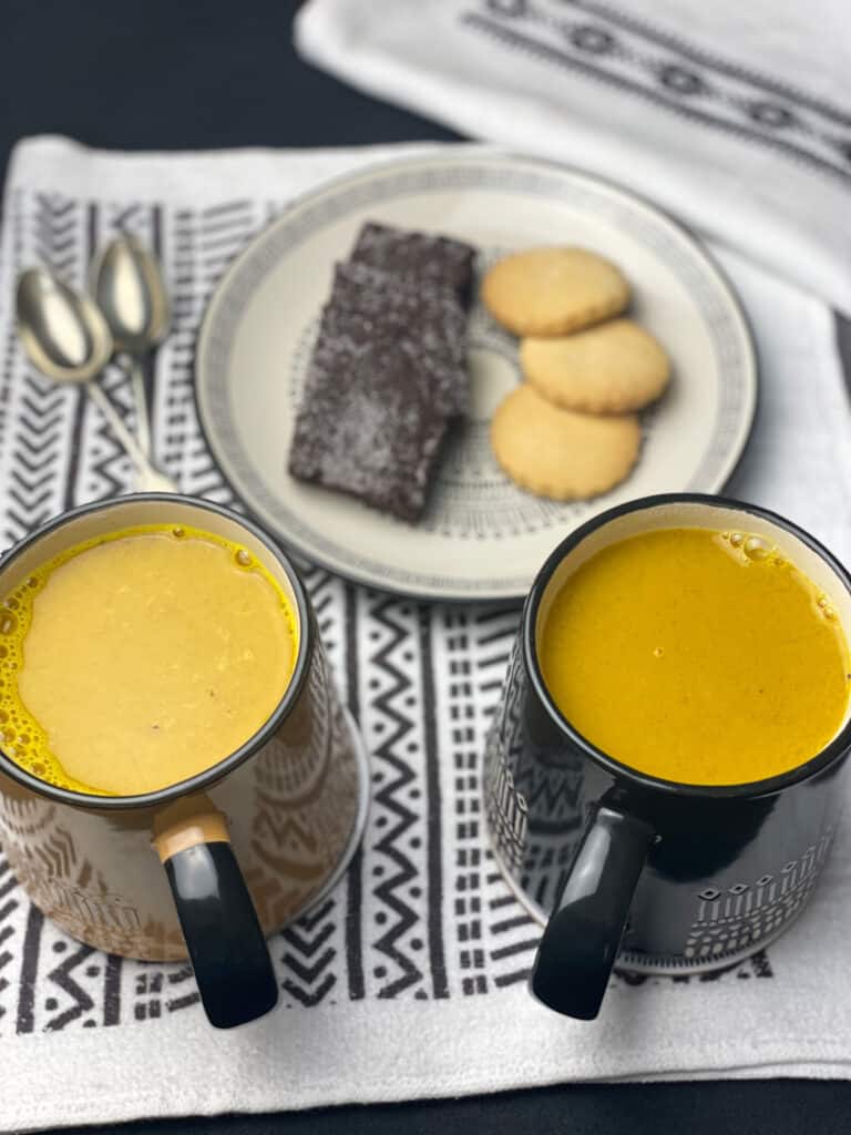 turmeric latte poured into 2 mugs, plate of cookies in front and 2 spoons, all on black and white pattern tea towel.