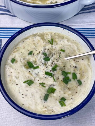 Chowder served in white bowl with blue rim and silver spoon, chopped chives scattered over.