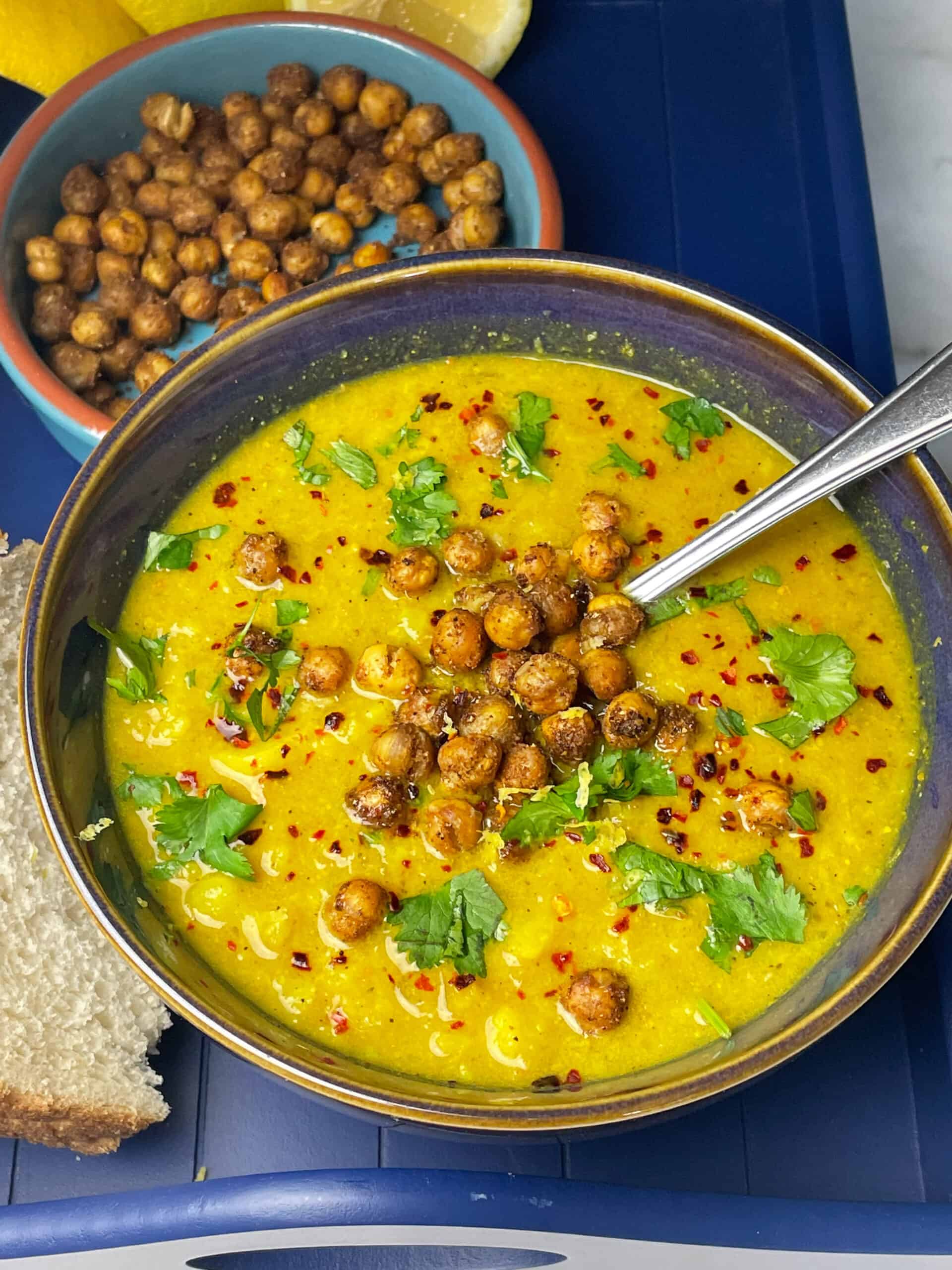 Bowl of curried cauliflower soup in blue bowl, slices of bread to side, roast chickpeas above, silver spoon and all sitting on blue tray.