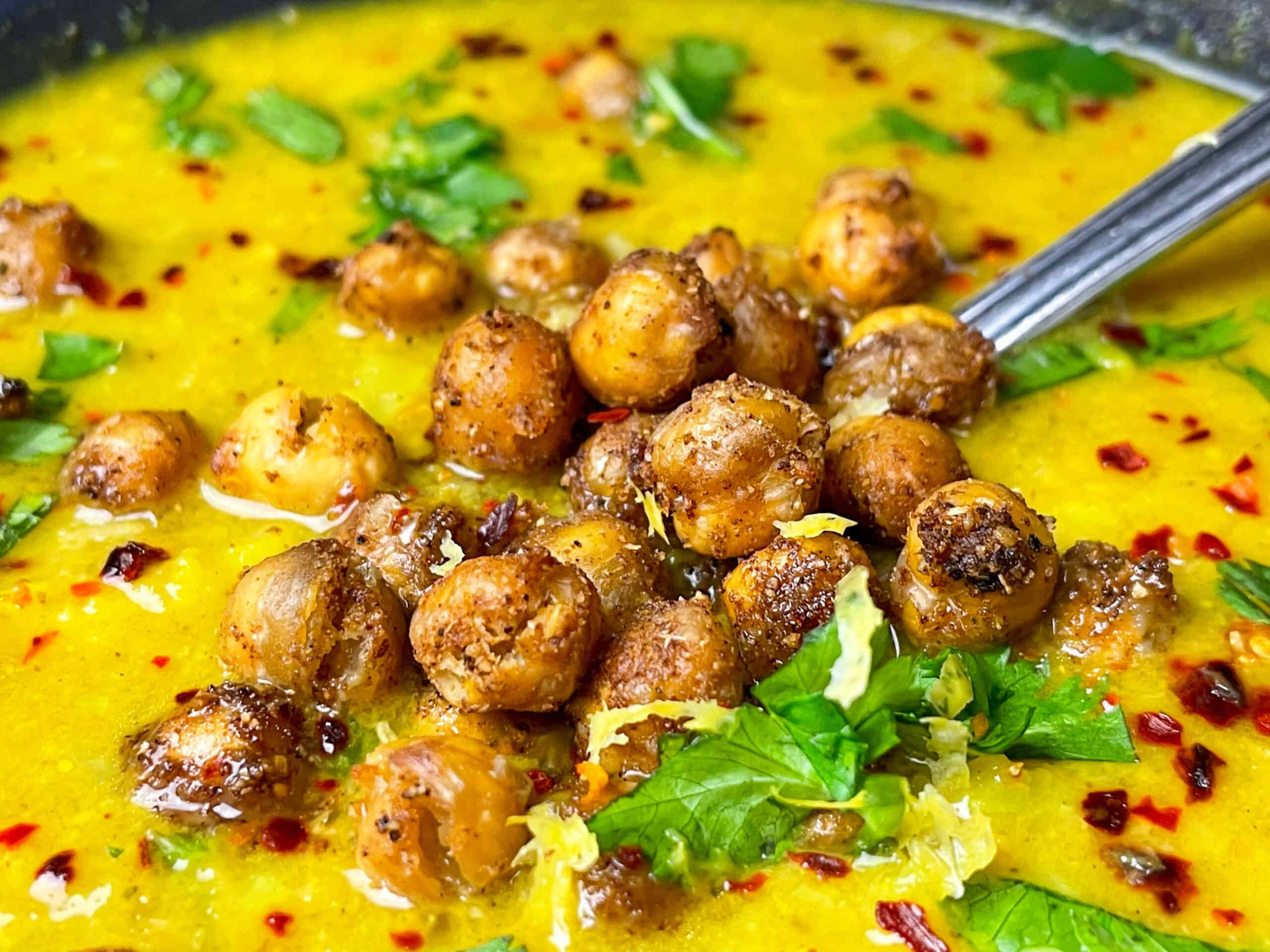 close up of vegan curried cauliflower soup with roast chickpeas on top and coriander, with silver spoon in soup.