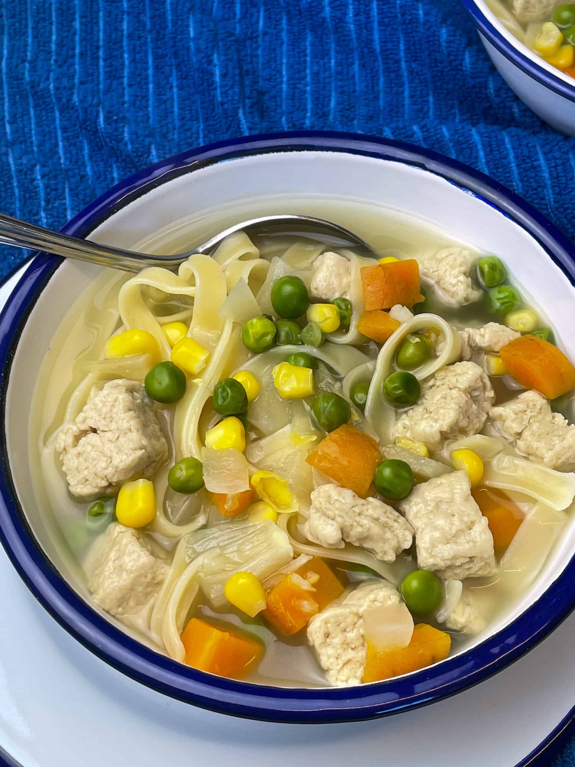 Bowl of chicken noodle soup with silver spoon and blue background.