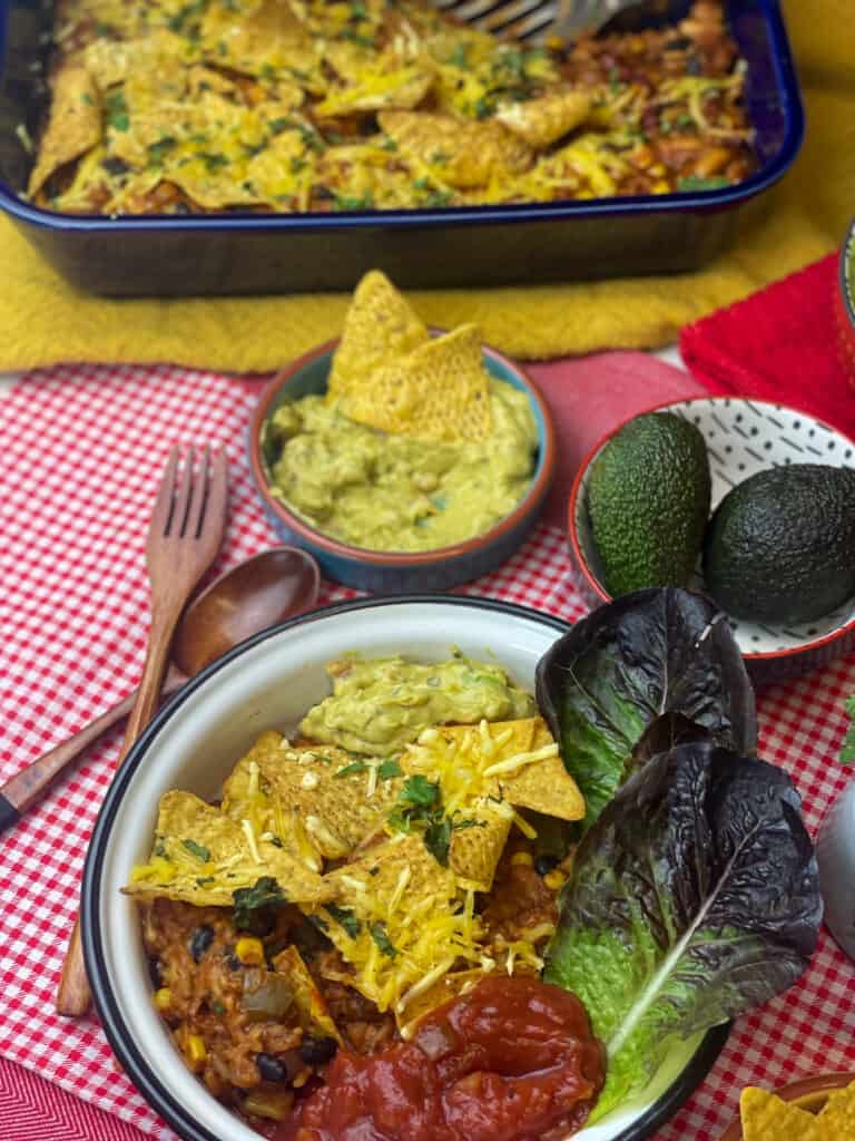 casserole served in bowl with salsa, guacamole and red lettuce leaves, small bowl of guacamole and avocados to side, and blue casserole dish in background, red and white check tablecloth and wooden fork.