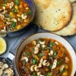 Two bowls of chickpea soup with small pan of cashews, lemon wedge to side, naan breads, all placed on grey tray.