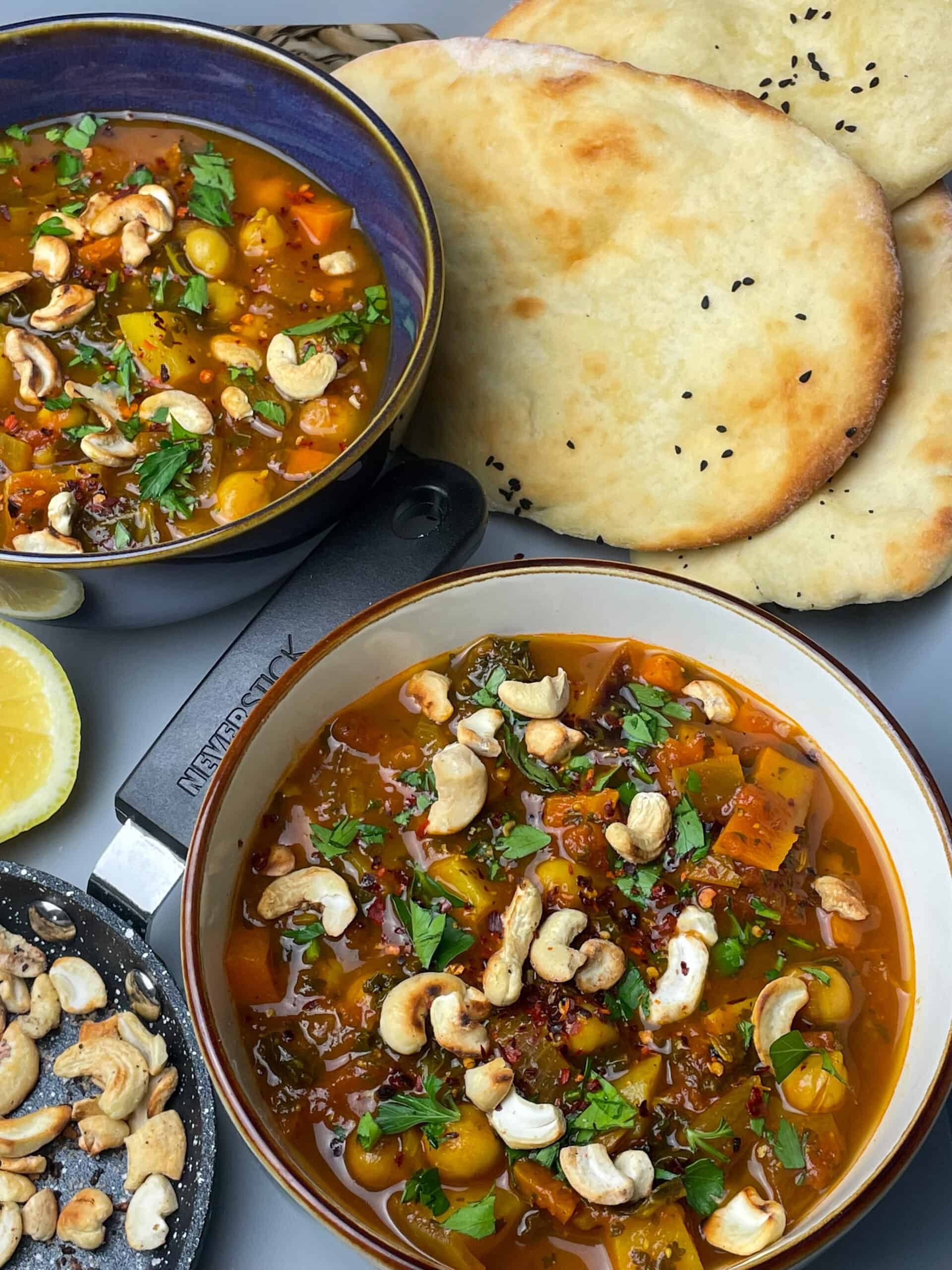 Two bowls of chickpea soup with small pan of cashews, lemon wedge to side, naan breads, all placed on grey tray.