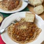 Two bowls of slow cooker green lentil spaghetti Bolognese, with sprinkle of nutritional yeast, wedge of her bread, silver fork and red check tea towel, featured image.