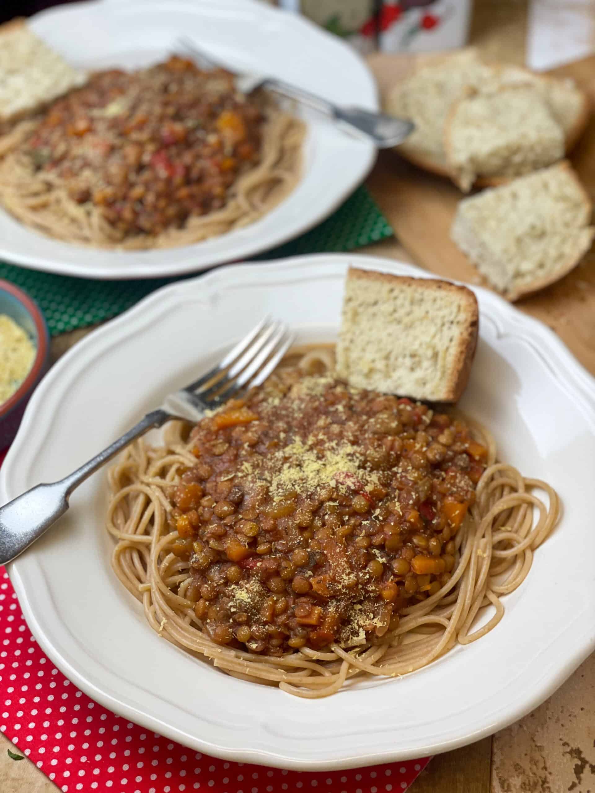 Two bowls of slow cooker green lentil spaghetti Bolognese, with sprinkle of nutritional yeast, wedge of her bread, silver fork and red check tea towel, featured image.
