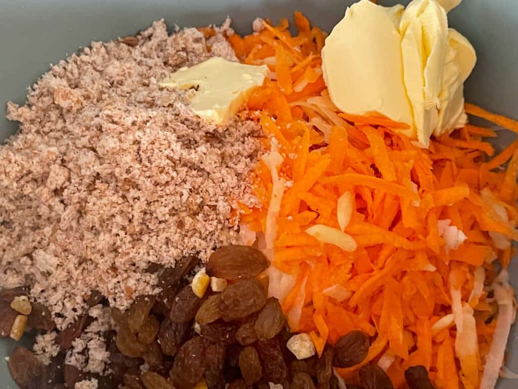 grated carrot and potato, margarine, dried fruit, breadcrumbs, flour, sugar and spices in green mixing bowl.
