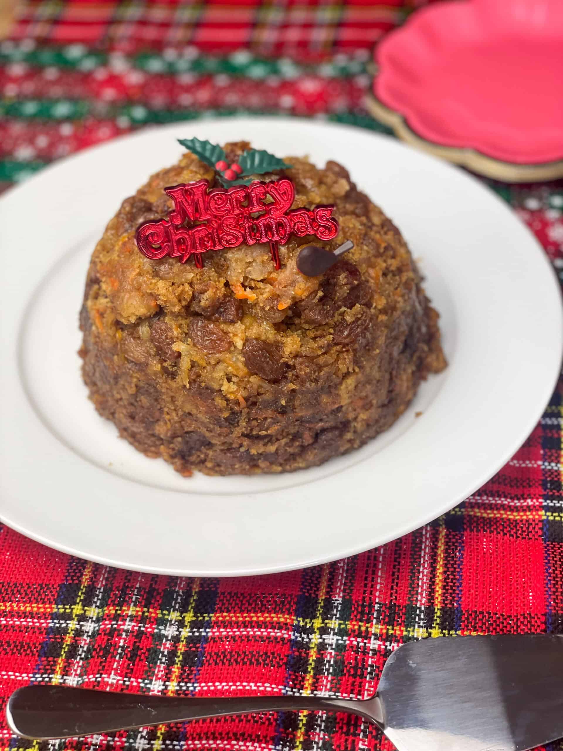 Christmas pudding sitting on white plate with red merry Christmas sign and little holly ornament, red tartan background.