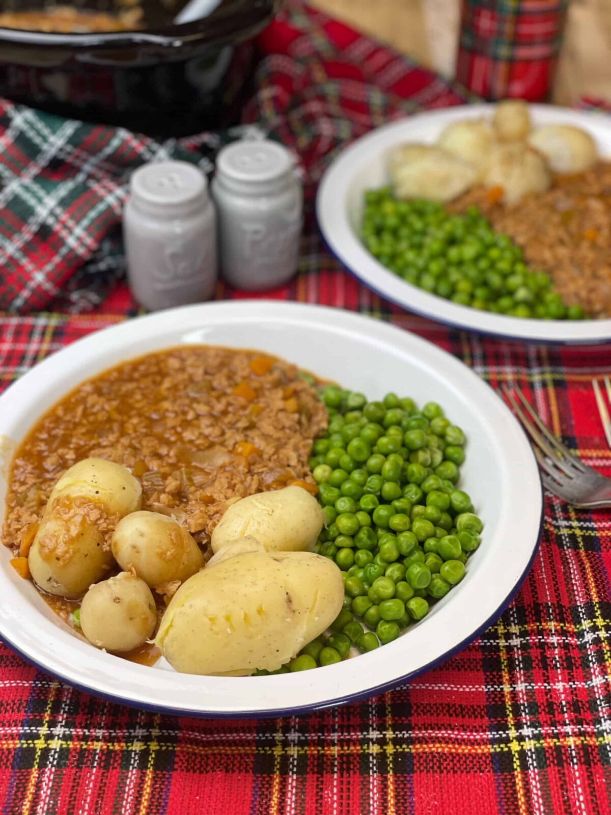 Two plates with vegan mince and tatties, peas, red tartan cloth underneath, blue salt and pepper shakers at the top, silver fork to side.
