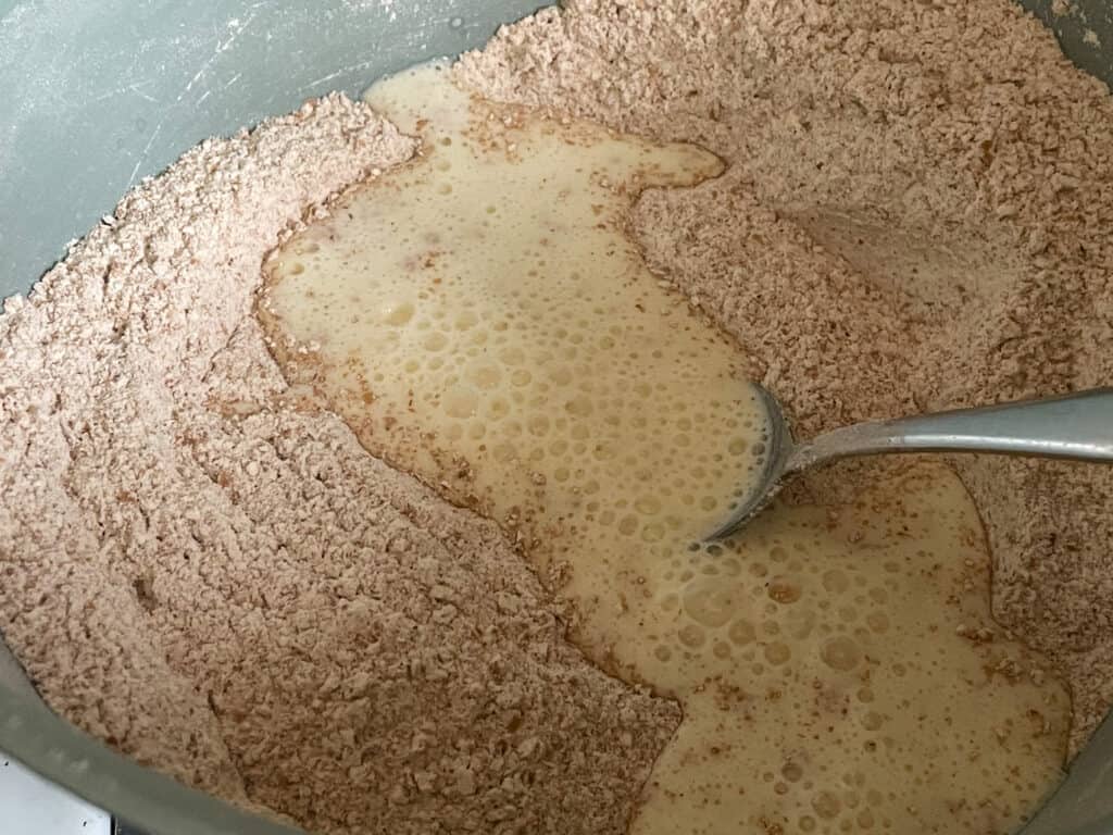 Wholemeal bread flour and bread ingredients in mixing bowl with soya buttermilk added, silver spoon in middle.