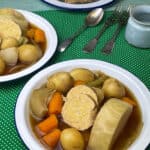 Two white enamel bowls of Irish vegan corn beef and cabbage stew, with plate of soda bread, cutlery and small cup, green polka dot background.