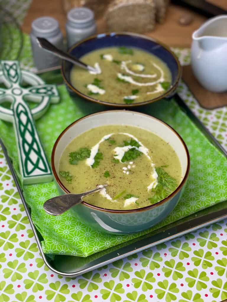 Two bowls of oatmeal leek broth on tray, with green flower tea towel, green and white celtic cross to side, white jug of cream to side, shamrock background,featured image.