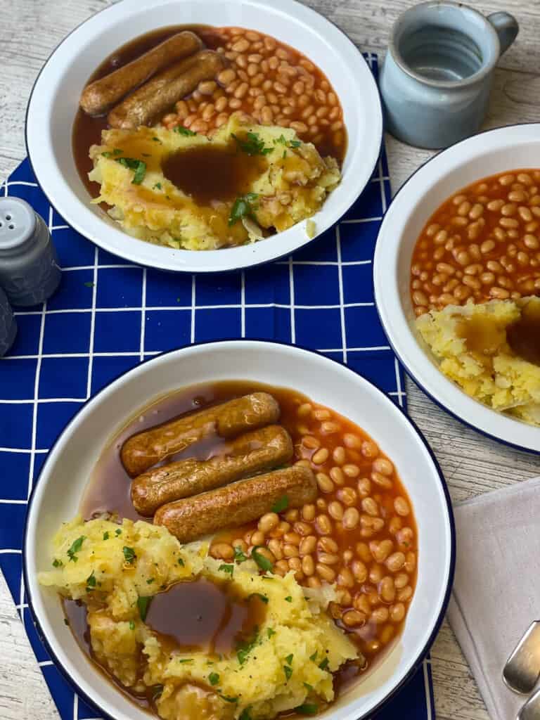 three bowls of champ with gravy poured over, vegan sausages to side and baked beans, blue with white check tea towel.