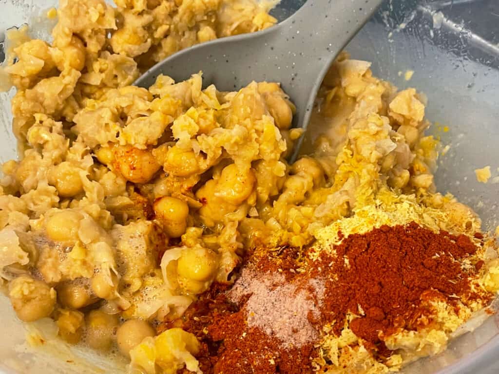 chickpeas mashed in bowl and spices added with grey spoon.