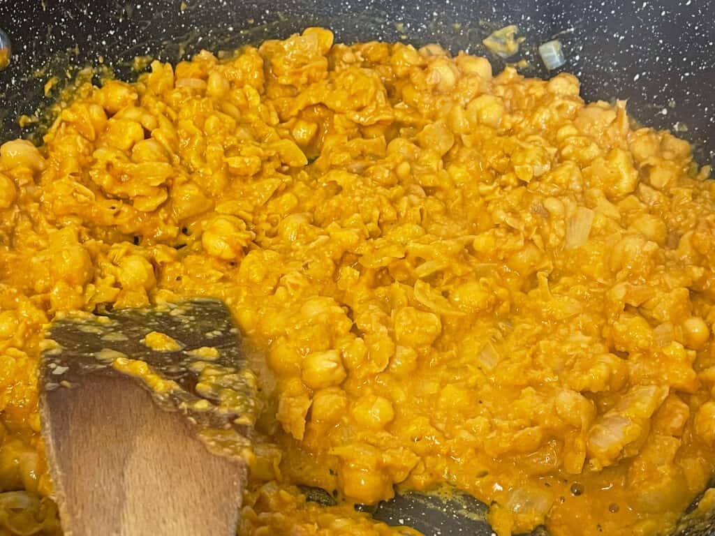 mashed chickpeas mixed with cooked onions in fry pan with spatula.